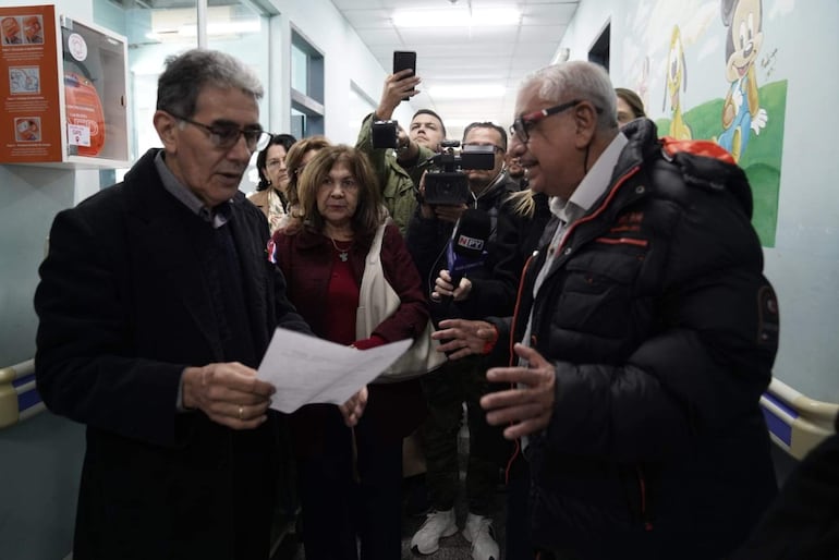 Isaías Fretes, con chaqueta oscura y cabello canoso, sostiene un documento mientras habla con otra persona en un pasillo institucional.