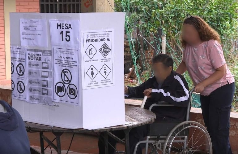 Mujer de cabello rizado en camiseta rosa asiste a hombre joven en silla de ruedas junto a una urna de votación en ambiente electoral.