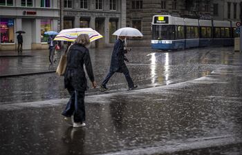 Personas caminan con paraguas mientras cruzan cerca de una conocida plaza en Zúrich (Suiza), en un día lluvioso.
