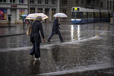 Personas caminan con paraguas mientras cruzan cerca de una conocida plaza en Zúrich (Suiza), en un día lluvioso.