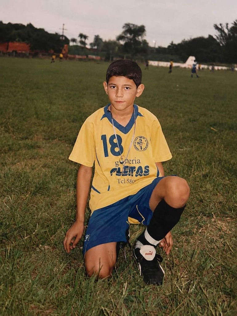 Niño con camiseta amarilla y azul, posando serio en el césped, mientras otros niños juegan al fútbol.