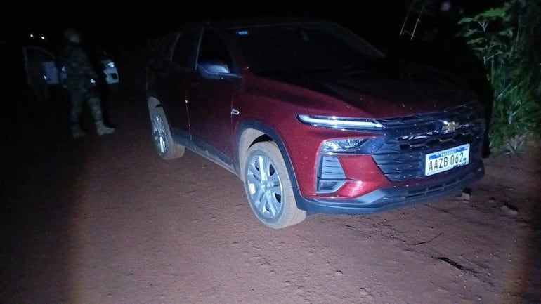 Uniformado militar en vigilancia junto a un auto rojo, en un camino de tierra, en un entorno rural. Iluminación tenue.