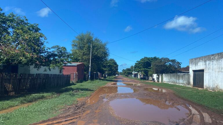 Calle del barrio San Roque, con casas a los lados, charcos y vegetación abundante. Cielo despejado y entorno tranquilo.