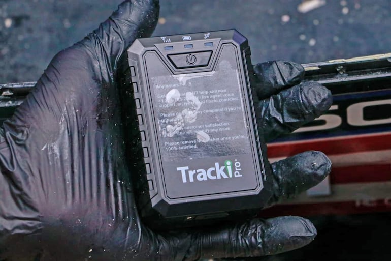 Dispositivo Tracki Pro sostenido por mano con guante negro, con pantalla mostrando información de soporte al cliente.