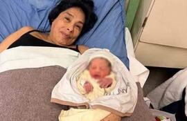 Mabel en cama de hospital, con camiseta negra, abrazando a su bebé David envuelto en manta clara, sonrisa serena.