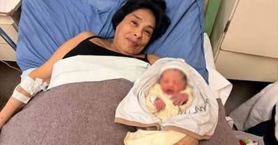 Mabel en cama de hospital, con camiseta negra, abrazando a su bebé David envuelto en manta clara, sonrisa serena.