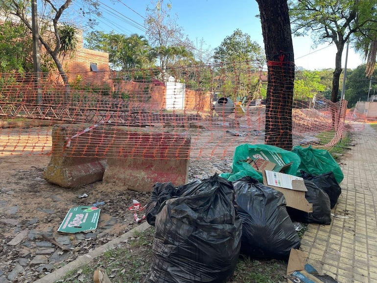 Entorno vacío con bolsas de basura y cartones, frente a obra en construcción delimitada por malla de seguridad naranja.