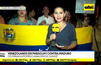 Presentadora de ABC en blusa oscura entrevista en la Plaza de la Democracia, rodeada de manifestantes con bandera de Venezuela y cartel de protesta.