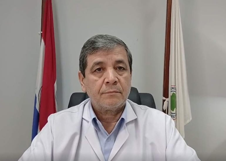 Hombre de mediana edad, con bata blanca y camisa clara, serio frente a la cámara, con banderas de Paraguay de fondo.