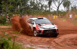 Vehículo Toyota en rojo y blanco, levantando polvo en un camino de tierra, mientras personas observan desde el fondo.