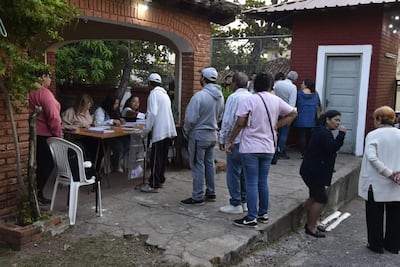 Grupo de personas en la entrada de un local de votación, con dos mujeres sentadas y un hombre de pie entregando documentación.