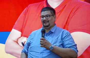 Hombre robusto, con camisa azul, sostiene un micrófono hablando a una audiencia. Fondo con colores rojo y azul.