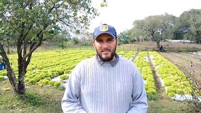 Hugo Del Puerto con suéter gris y gorra, frente a un extenso campo de lechugas, acompañado de otros trabajadores en el fondo.
