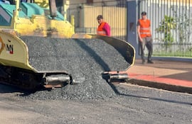 Dos trabajadores en chalecos naranjas, uno con pala y el otro con carretilla, observados por un tercero en un entorno de construcción.