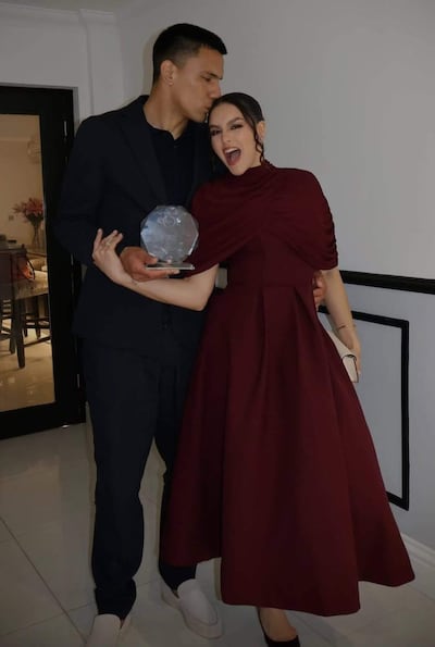Diego Gómez con traje oscuro besa a Geraldine Villagra, elegante en vestido burdeos, sosteniendo un trofeo y sonriendo feliz.