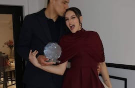 Diego Gómez con traje oscuro besa a Geraldine Villagra, elegante en vestido burdeos, sosteniendo un trofeo y sonriendo feliz.