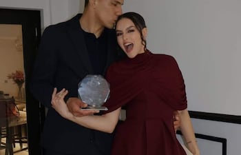 Diego Gómez con traje oscuro besa a Geraldine Villagra, elegante en vestido burdeos, sosteniendo un trofeo y sonriendo feliz.