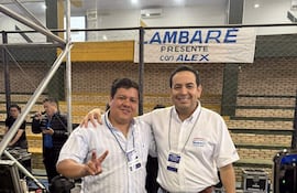 IZA AMBARÉ en camisa de cuadros y Alex en camisa blanca sonríen, rodeados de un ambiente de auditorio con gradas.