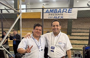 IZA AMBARÉ en camisa de cuadros y Alex en camisa blanca sonríen, rodeados de un ambiente de auditorio con gradas.