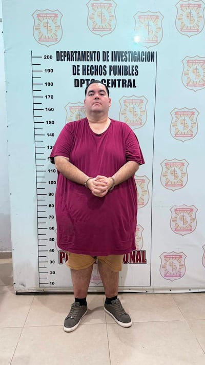 Hombre con camiseta burdeos y pantalones cortos claros, de pie con manos entrelazadas, fondo con emblema de la Policía Nacional.