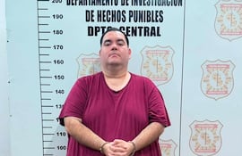 Hombre con camiseta burdeos y pantalones cortos claros, de pie con manos entrelazadas, fondo con emblema de la Policía Nacional.