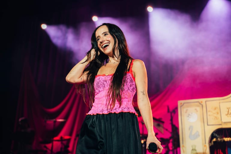 Mujer de pie en escenario con micrófono, sonriente, blusa rosa y pantalones oscuros, fondo festivo iluminado.