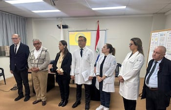 Grupo de siete personas en conferencia, tres con batas blancas y otros en traje, con bandera de Paraguay de fondo.