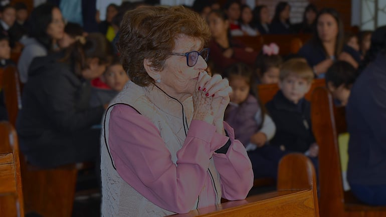 Mujer mayor con cabello rizado y gafas, en oración, rodeada de niños y adultos en un banco de madera en un recinto religioso.