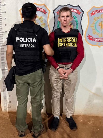 Agente de policía de espaldas con camiseta negra y hombre sereno en camiseta roja y chaleco negro etiquetado como 'INTERPOL DETENIDO'.