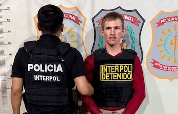 Agente de policía de espaldas con camiseta negra y hombre sereno en camiseta roja y chaleco negro etiquetado como 'INTERPOL DETENIDO'.