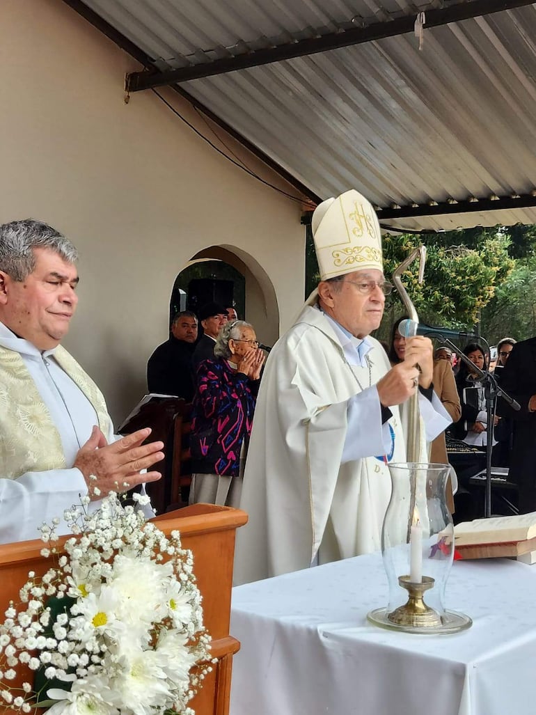 Sacerdote de mitra y vestimenta religiosa habla al micrófono, mientras otro hombre observa en un evento al aire libre.