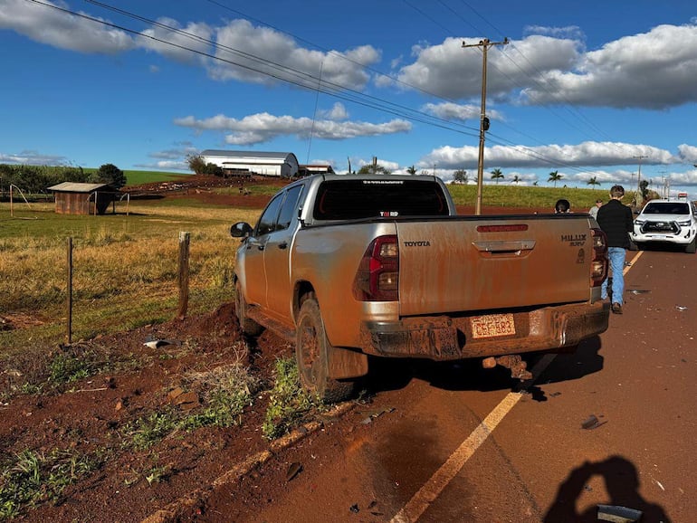 Dos personas observan una Toyota Hilux sucia en un camino rural, mientras un vehículo blanco se detiene cerca.