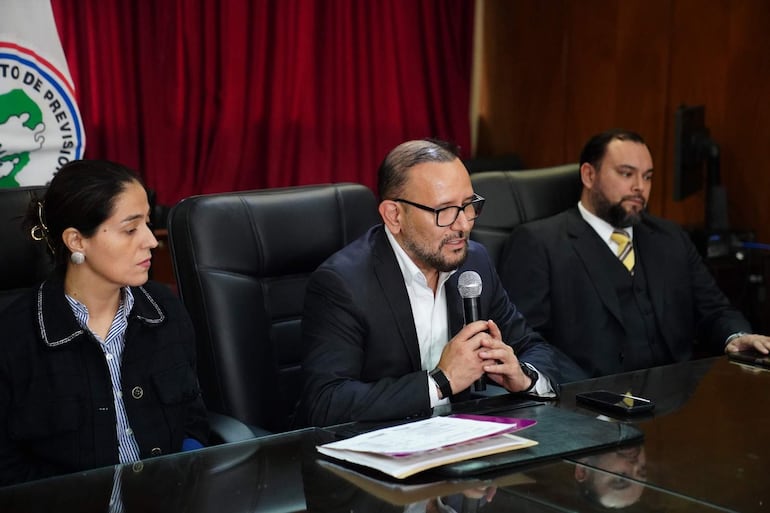 Pablo José Morinigo Cáceres habla con micrófono, acompañado de hombre y mujer atentos, en salón con cortina burdeos.