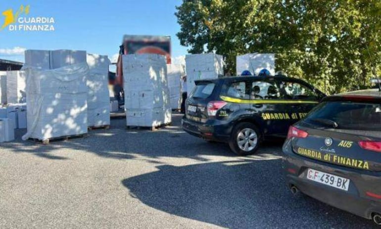 Autos patrulleros de la Guardia di Finanza en un área industrial con pallets de cajas blancas apiladas detrás.