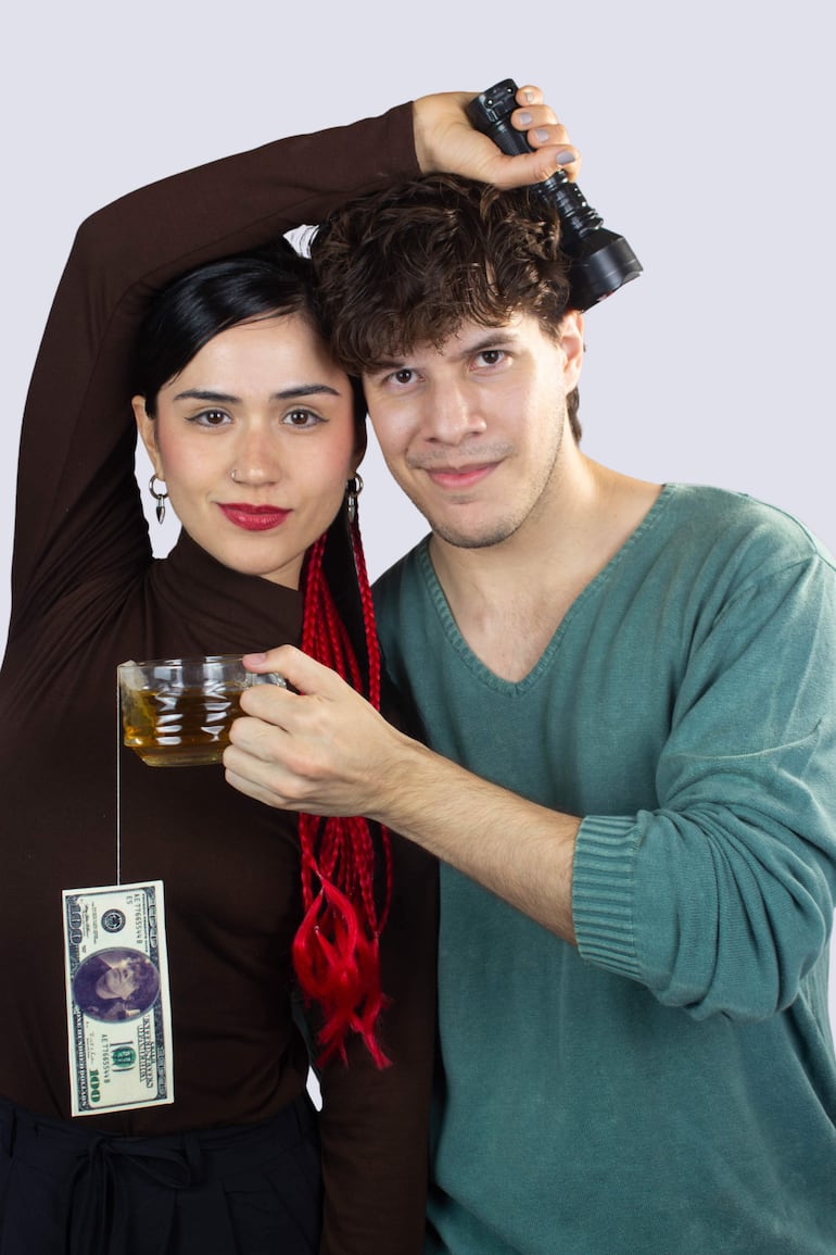 Mujer de trenzas rojas y prenda marrón levanta un vaso amarillo, mientras un hombre con camiseta verde señala con una linterna.