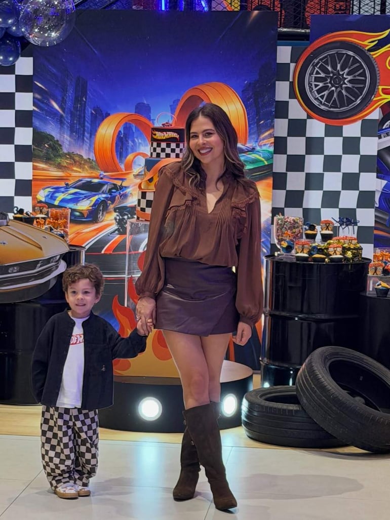 Mujer sonriente con blusa marrón y falda corta de cuero, sostiene la mano de un niño en camiseta blanca y pantalones de cuadros, rodeados de decoraciones coloridas.