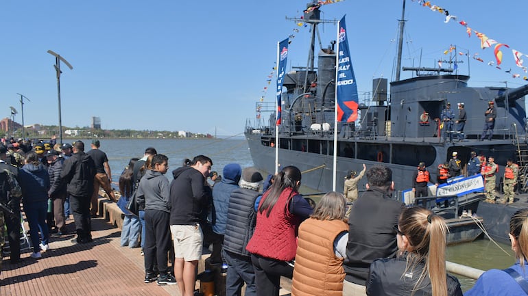 Grupo de personas en abrigos mira un barco militar adornado con banderas en un muelle de Encarnación.