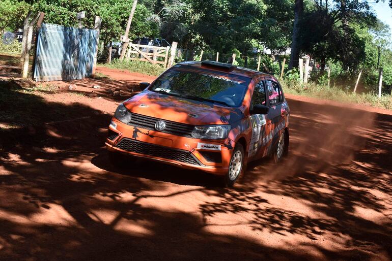 Vehículo de competición de rally, color naranja, levantando polvo en un camino rural entre vegetación.