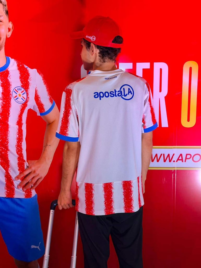 Dos jóvenes, uno de pie con camiseta a rayas rojas y blancas, sosteniendo un carrito, en fondo rojo con texto promocional.