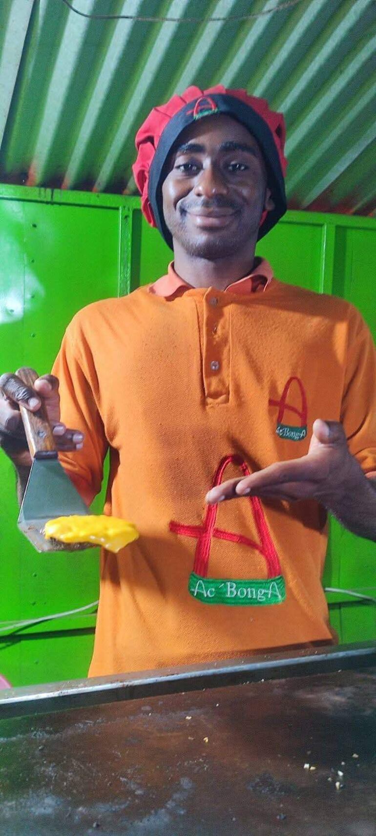 Alfredo Bonga sonriente, vistiendo camiseta naranja y sosteniendo una espátula, en un ambiente de cocina colorido.