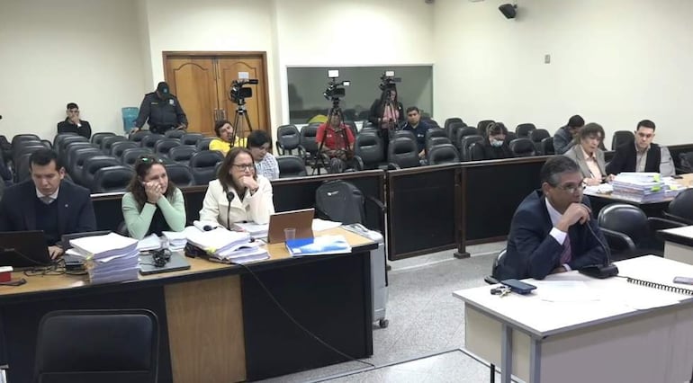 Hermann Weisensee en sala de audiencias, rodeado de personas concentradas, con una mujer de blusa verde pensativa junto a otra mujer de abrigo blanco.
