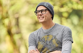 Daniel H. Wilson sonríe con gorro gris y camiseta gris, rodeado de vegetación difusa en un ambiente relajado.