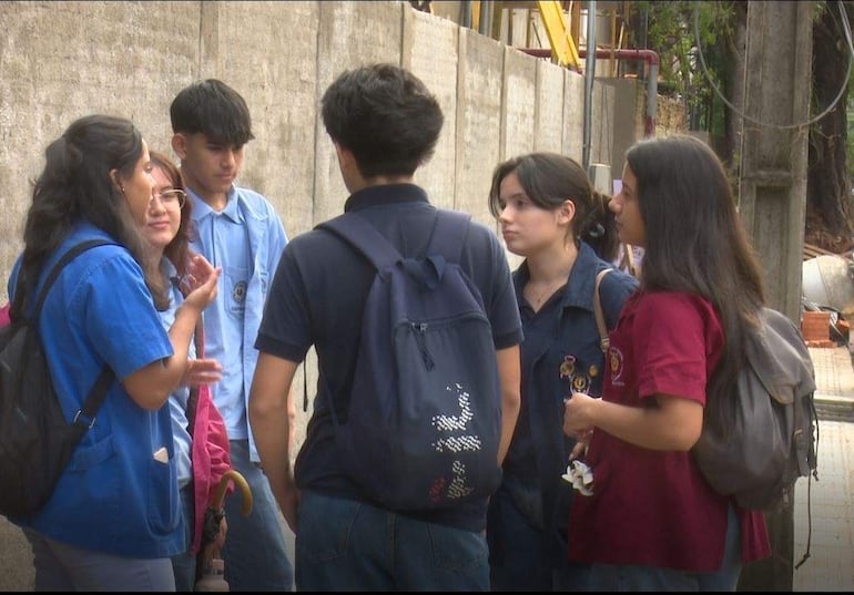 Grupo de seis jóvenes conversando al aire libre, una mujer con cabello largo y oscuro habla animadamente mientras los demás la escuchan.