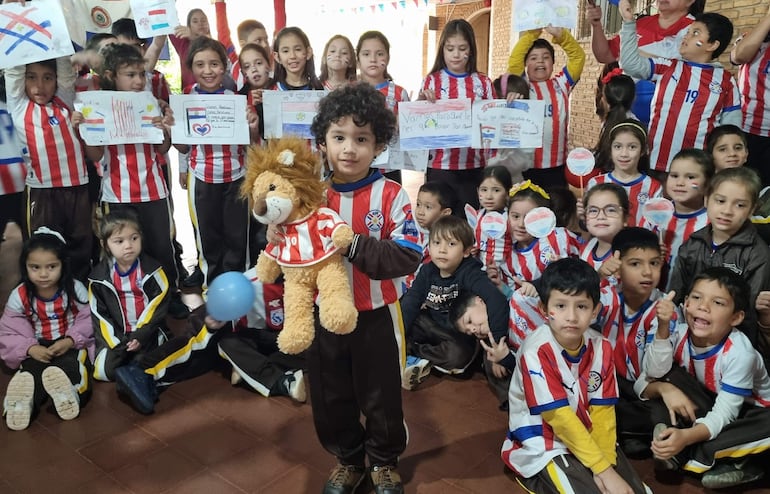 Niños vestidos con camisetas rojas y blancas, sosteniendo carteles y un peluche de león, en un ambiente festivo lleno de alegría.
