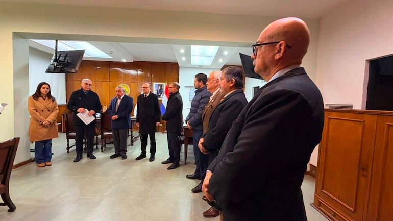 Isaías Fretes en abrigo negro habla durante una reunión, rodeado de personas vestidas formalmente en un ambiente institucional.
