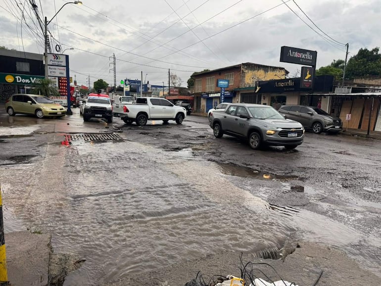 Motociclista detenido junto a vehículos en calle mojada, con baches visibles y letreros de comercios alrededor, en un entorno lluvioso.