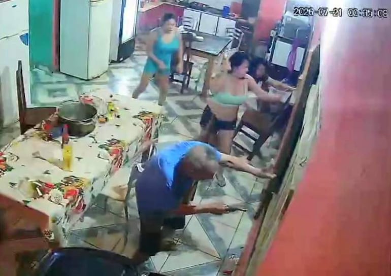 Hombre de edad avanzada agachado cerca de una puerta, rodeado de mujeres en una cocina con mantel colorido.