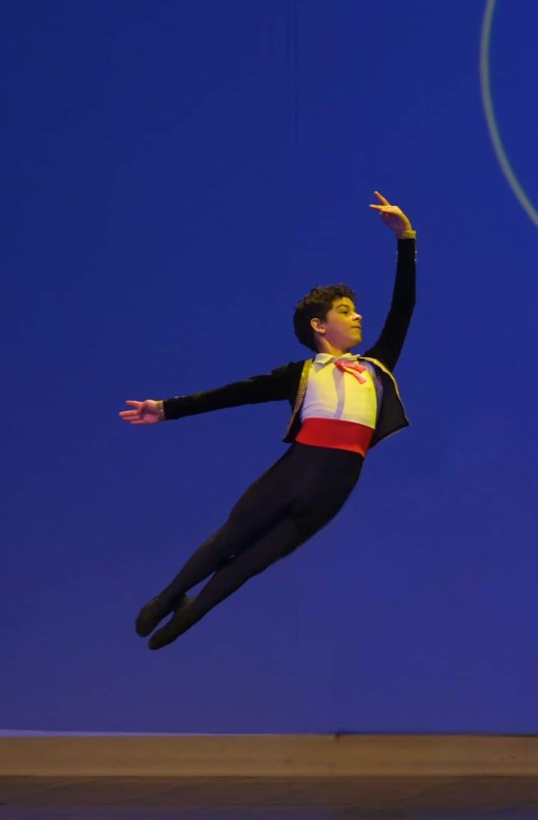 Niño en salto con chaleco negro y camisa blanca, cinta roja en la cintura, rodeado de fondo azul en un contexto de danza.