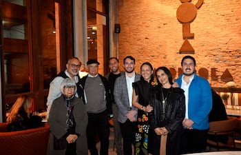 Una mujer en vestido negro se destaca entre ocho personas sonrientes en un interior acogedor con decoración de ladrillo expuesto.