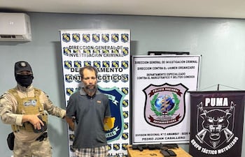 Agente policial con uniforme y máscara sostiene a Luis Carlos De Jesús, vestido con camiseta gris y pantalones cortos. Oficina bien iluminada.